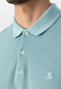 Cou du homme et haut de poitrine portant un polo bleu clair texturé avec deux boutons et des lettres blanches brodées "MOP" sur le côté gauche.