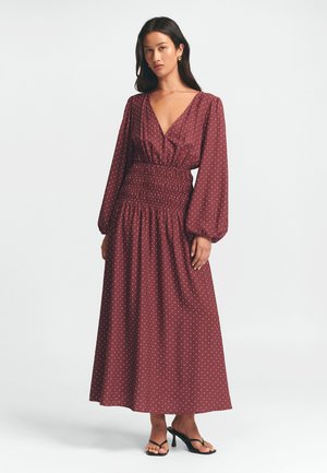 Burgundin täplikkää maxi-mekko syvällä V-kaula-aukolla, pitkillä hihoilla ja kuminauhakiristetyllä vyötäröllä, valmistettu kevyestä kankaasta, yhdistetty mustiin sandaaleihin.