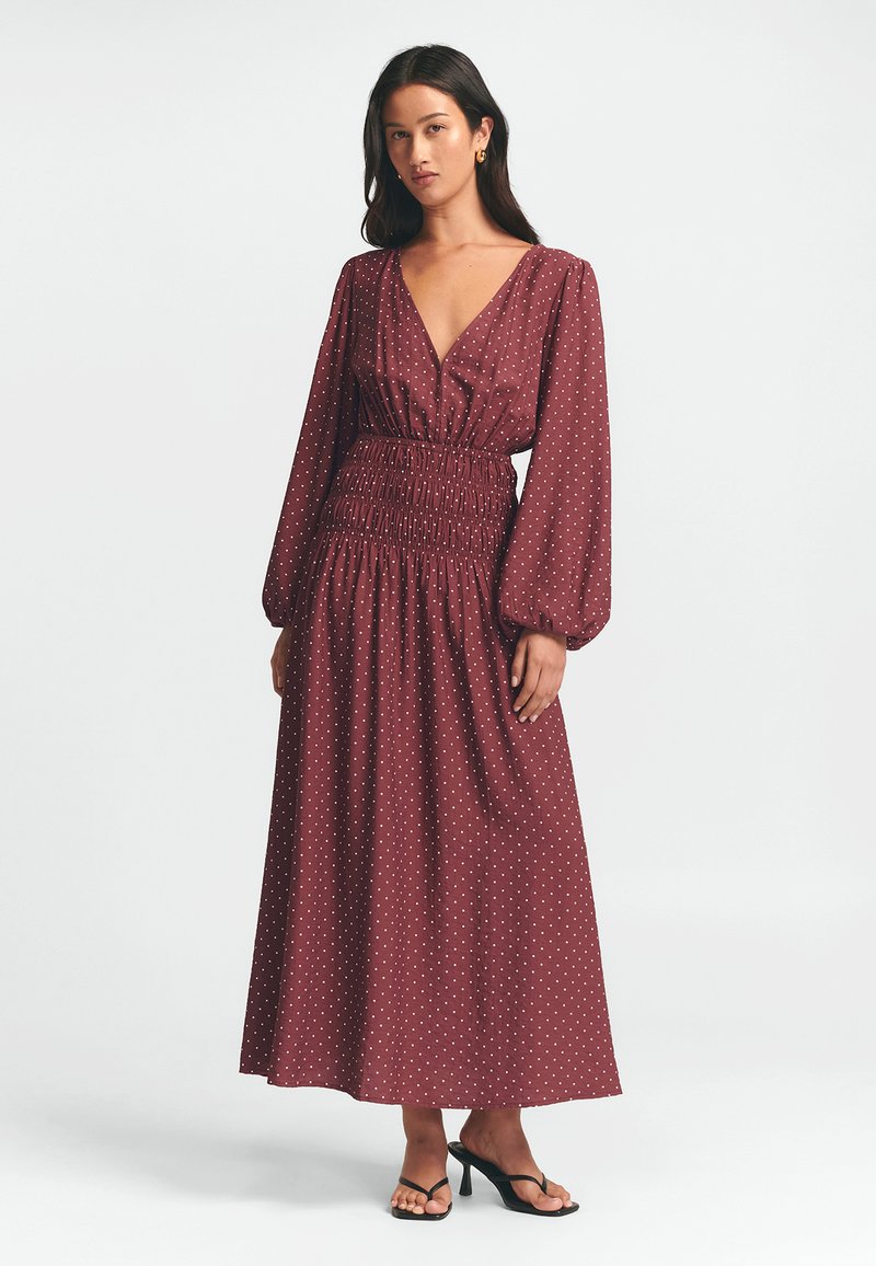 Burgundská maxi šaty s puntíky, s hlubokým V-výstřihem, dlouhými rukávy a smockovaným pasem, vyrobené z lehkého materiálu, v kombinaci s černými sandály.
