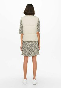 Gilet rembourré beige avec un design matelassé, porté sur une robe à motifs. L'ensemble présente des manches courtes et des baskets blanches.