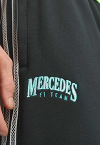 Schwarze Sporthose mit weißen Netzeinsätzen an den Seiten, mit türkisfarbenem besticktem Schriftzug "MERCEDES F1 TEAM." Glatter Stofftextur.