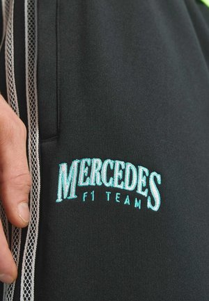 Pantalones deportivos negros con paneles laterales de malla blanca, con texto bordado en turquesa que dice "MERCEDES F1 TEAM." Textura de tela suave.