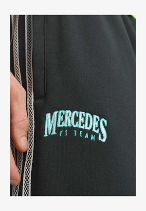 Čierne športové nohavice s bielymi sieťovanými bočnými panelmi, s tyrkysovým vyšívaným textom "MERCEDES F1 TEAM." Hladká textúra látky.