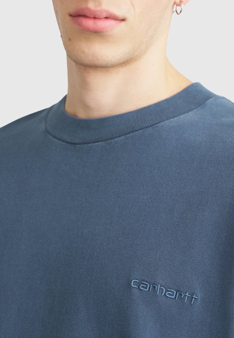 Detailní záběr osoby, která má na sobě modrou mikinu Carhartt a stříbrný kruhový náušník v levém uchu.