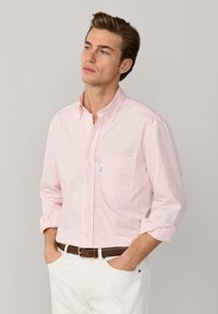 Chemise rose pâle à boutons avec rayures blanches, manches longues retroussées, une poche poitrine, tissu en coton. Portée avec un pantalon blanc et une ceinture marron.