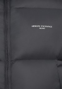 Jachetă neagră cu umplutură, cu închidere prin fermoar, având textul alb "ARMANI EXCHANGE MILANO" imprimat aproape de guler. Textură netedă.