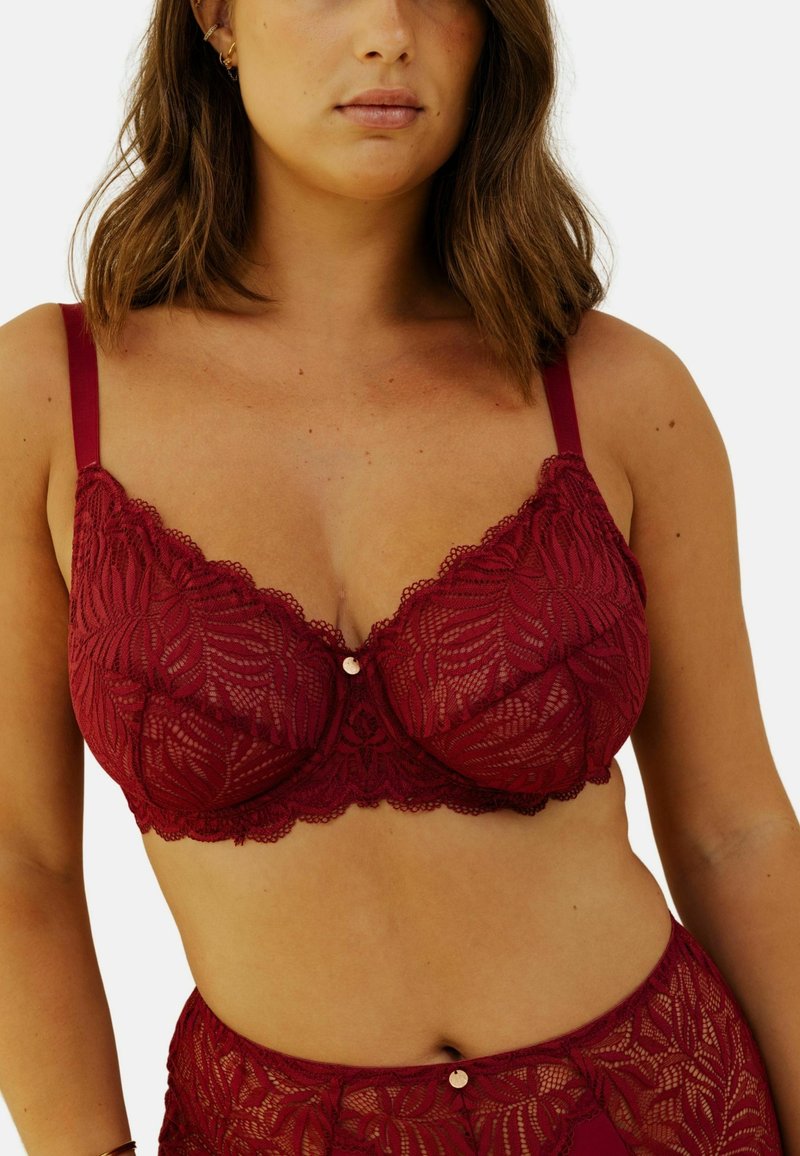 Soutien-gorge en dentelle bordeaux avec motif floral, bonnets transparents, larges bretelles et un petit accent métallique décoratif au centre. Texture douce.
