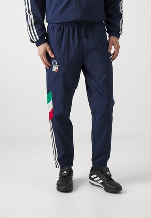 Tuta navy con strisce bianche sulle maniche, con dettagli della bandiera italiana in verde, bianco e rosso sui pantaloni. Vita con coulisse, tessuto leggero.