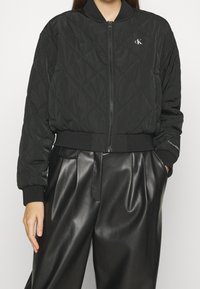 Czarna pikowana kurtka bomberka z zapięciem na zamek, żebrowanymi mankietami i dołem. Posiada małe logo Calvin Klein na piersi. Noszona z czarnymi skórzanymi spodniami.