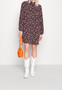 Kleid mit floralem Muster in Schwarz, das pinke und weiße Blumen zeigt, lange Ärmel und einen Kragen hat. Kombiniert mit einer orangefarbenen Handtasche und weißen Cowboystiefeln.