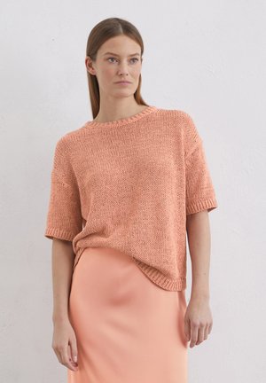 Maglione lavorato a maglia a maniche corte in corallo, con una vestibilità ampia e un tessuto lavorato. Indossato sopra una gonna liscia di colore rosa pallido.