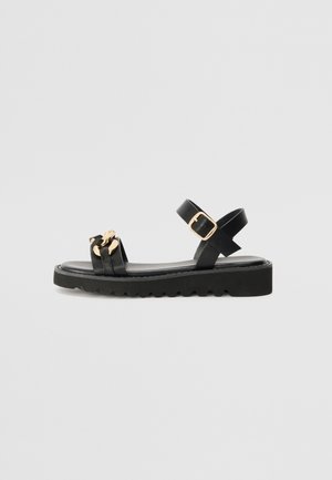 Black sandal featuring wide straps, a gold accent, and a thick, ridged sole. Adjustable ankle strap with a buckle detail. Smooth texture.