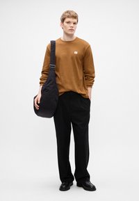 Καφέ μακρυμάνικη υφασμάτινη μπλούζα με μικρό λογότυπο, μαύρη τσάντα ώμου με λεία επιφάνεια, χαλαρούς μαύρους παντελόνες και μαύρα παπούτσια.