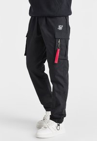 Svarta cargobyxor med en strukturerad yta, med sidofickor och en blixtlåsdetaill med en röd dragflik. I kombination med vita sneakers.