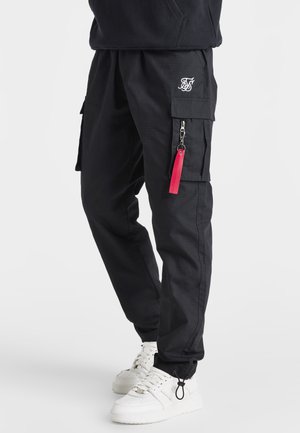 Schwarze Cargo-Hosen mit strukturierter Oberfläche, ausgestattet mit Seitentaschen und einem Reißverschlussdetail mit einem roten Zugband. Kombiniert mit weißen Sneakers.