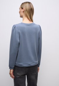 Sininen huppari pitkillä hihoilla, pyöreällä kaula-aukolla ja puffattuja olkapäitä. Valmistettu pehmeästä kankaasta, jossa on rento istuvuus ja ribbireuna.