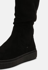 Bottine en daim noir avec une semelle en caoutchouc nervurée, présentant des détails de couture courbés et un design ajusté. Texture lisse partout.