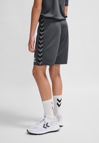 Grijze sportshorts met zwarte chevron-zijstrepen, gecombineerd met witte sneakers en sokken met zwarte chevronpatronen.