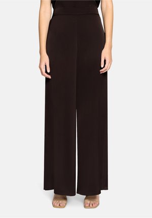 Frau trägt hoch taillierte, weit geschnittene schwarze Hosen kombiniert mit beigefarbenen Sandalen mit offenen Zehen, steht vor einem hellgrauen Hintergrund.