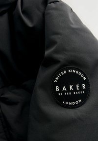 Détail d'une veste en tissu noir avec un patch logo rond cousu qui indique "BAKER par TED BAKER LONDRES" et "ROYAUME-UNI."