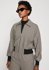Jacke mit Zickzackmuster in Schwarz und Weiß, mit Reißverschluss vorne, weitem Kragen und gerippten Bündchen. Kombiniert mit einem schwarzen Oberteil.
