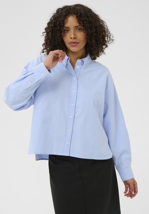 Femme aux cheveux bouclés portant une chemise bleu clair boutonnée et une jupe noire, tenant le col de sa chemise d'une main.