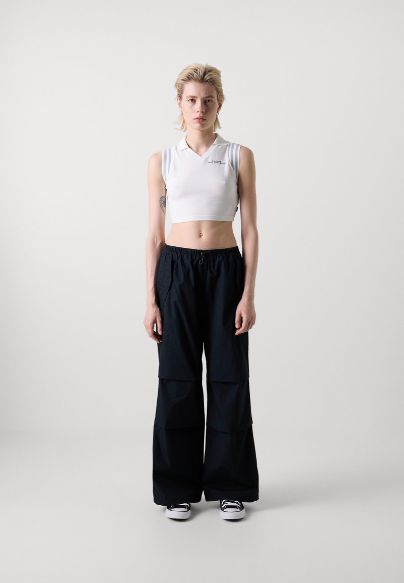 T-shirt polo blanc court avec un col en V, associé à un pantalon cargo noir ample avec des poches, stylisé avec des baskets noires et blanches.