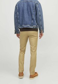 Giubbotto di denim, lavaggio blu, con dettagli in bottone; top marrone sotto; chinos slim-fit color beige; calzature marroni con suole chiare.