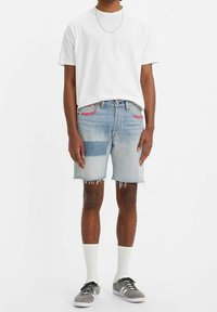 Ljust blå denimshorts med fransade hörn, prydda med kontrasterande lappar och rosa sömmar, i kombination med en enkel vit t-shirt och grå sneakers.