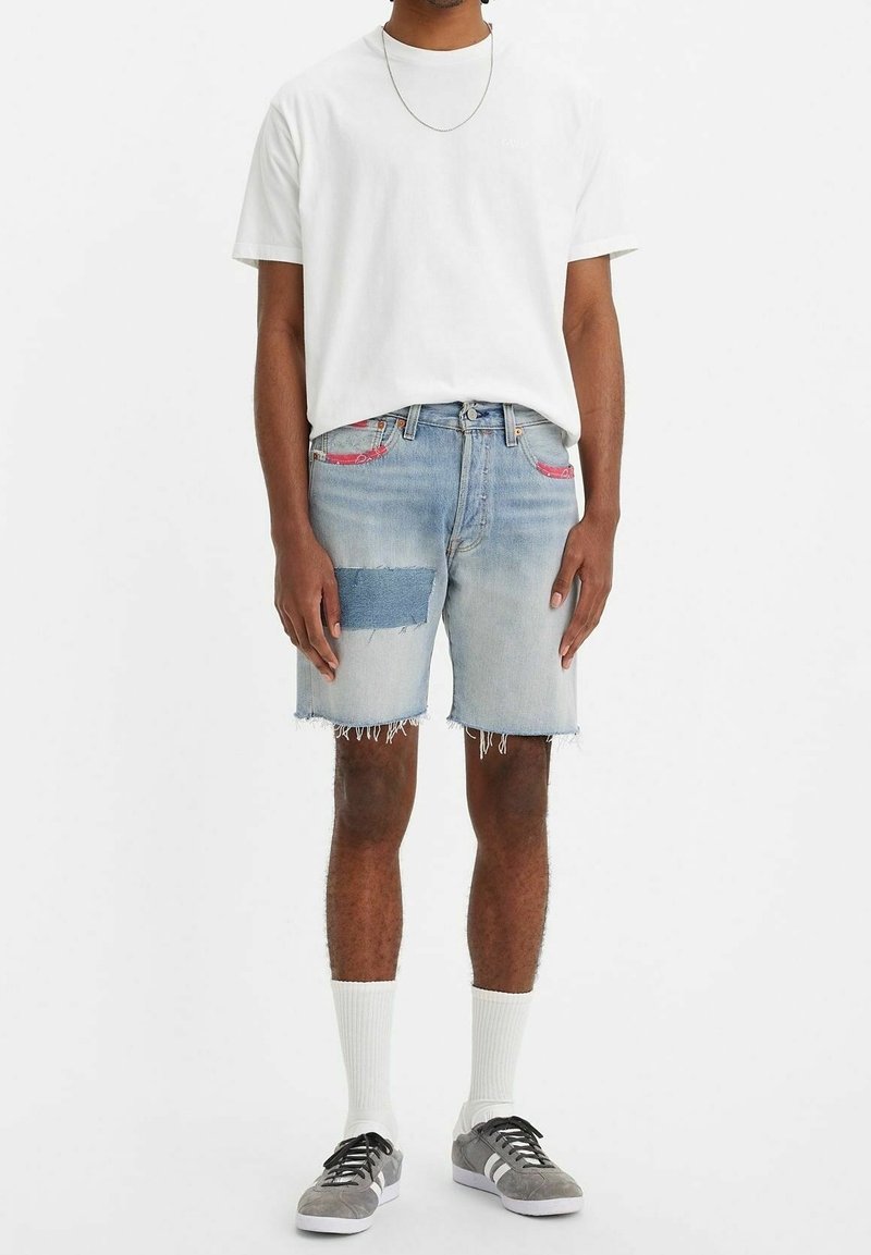 Ljust blå denimshorts med fransade hörn, prydda med kontrasterande lappar och rosa sömmar, i kombination med en enkel vit t-shirt och grå sneakers.