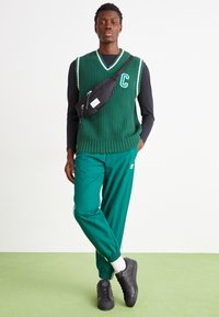 Zelený pletený svetr s bílým lemem a písmenem 'C', spárovaný se zelenými teplákovými kalhotami a černými teniskami, s černou crossbody taškou.