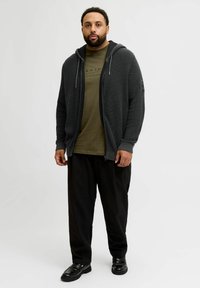 Sweat à zip gris foncé avec un motif texturé, superposé sur un t-shirt vert olive ; associé à un pantalon noir ample et des chaussures noires.