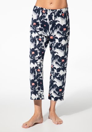Frau trägt weiße, kurz geschnittene Hose mit marineblauem und korallenfarbenem Blumenmuster, steht barfuß vor weißem Hintergrund.