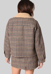 Bruine geblokte jas met een beige, pluchen kraag. Heeft een losse pasvorm, verlaagde schouders en een middellange snit met een uitsparing aan de achterkant.