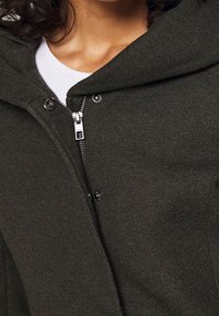 Sudadera verde oscuro con cremallera, botones a presión y una textura suave. Presenta una capucha amplia y un estilo superpuesto sobre una camiseta blanca.