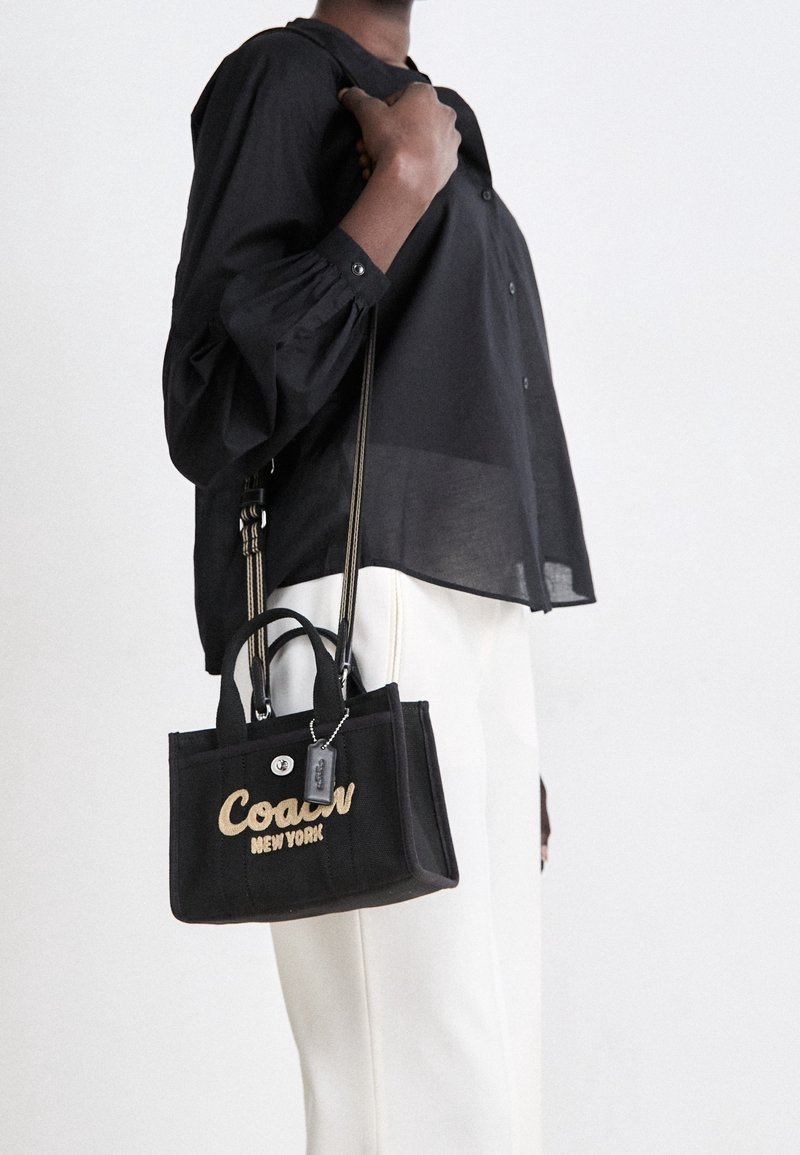 Personne portant une chemise noire à manches longues et un pantalon blanc tenant un sac à main noir Coach New York avec poignées doubles et bandoulière.