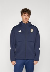 Navy-blauer Zip-Up-Jacke aus weichem Material, mit weißen Adidas-Streifen, Real Madrid-Logo auf der Brust und hohem Kragen.