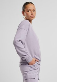 Felpa lilla con maniche lunghe, vestibilità comoda, polsini a costine e tasche laterali. Materiale morbido con una texture liscia.