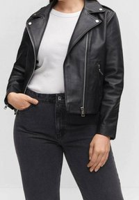 Personne portant une veste en cuir noir de motard par-dessus une chemise blanche avec un jean taille haute foncé, une main dans la poche et l’autre reposant à côté.