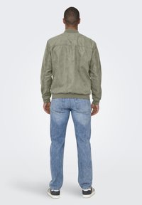 Olivgrüne Wildleder-Bomberjacke mit gerippten Bündchen, getragen über hellblauen Jeans. Die Jacke hat ein einfaches, klassisches Design.