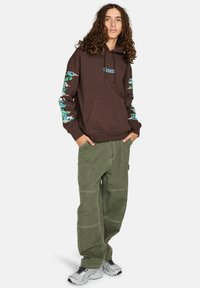 Bruine hoodie met grafische prints op de mouwen, voorzak. Gecombineerd met olijfgroene baggy broek en grijze sneakers.
