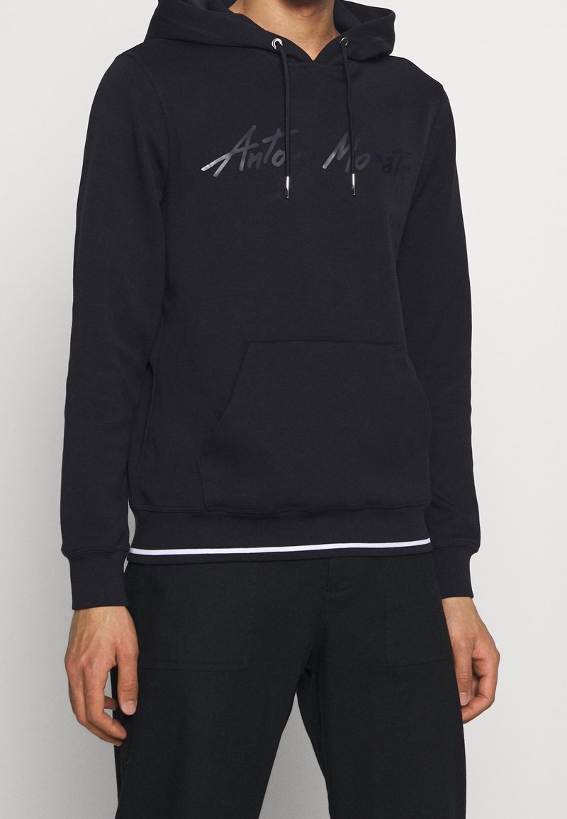 Svart hoodie med en framföre känguruficka, dragsnodd på huven och silverfärgad logotext. Har ribbade muddar och nederkant med ett vitt accentband.