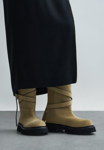 Karmelowe zamszowe kozaki do kolan z okrągłymi paskami i grubą czarną podeszwą, noszone pod czarną spódnicą z bocznym zamkiem.