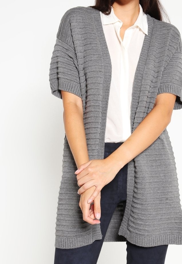 Grå stickad cardigan med korta ärmar, ribbat mönster och öppen framsida. Kombinerad med en vit skjorta med krage och mörka byxor.