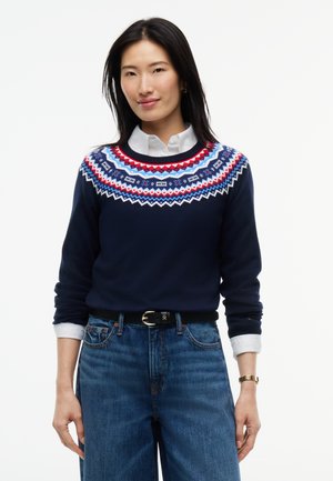 Mujer de pie vestida con un suéter azul marino con canesú estampado en rojo, blanco y azul, camisa blanca, vaqueros azules de pierna ancha, cinturón negro y zapatos negros.