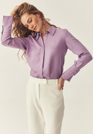 Kvinde med langt hår iført lilla skjorte med knapper og hvide bukser med høj talje, poserer med den ene hånd i håret og lukkede øjne.