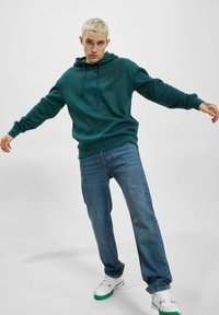 Blågrønn hettegenser med justerbare snorer, avslappet passform og ribbestrikkede mansjetter, kombinert med lyseblå jeans og hvite joggesko med grønne detaljer.