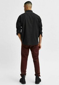 Camisa negra de manga larga con puños enrollados, combinada con pantalones burdeos y botines negros, vista desde atrás. Diseño simple y moderno.