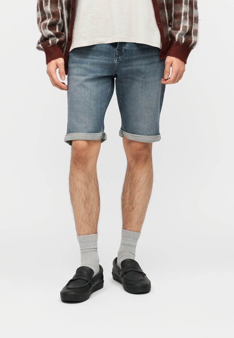 Άνδρας που φοράει τζιν σορτς γυρισμένα, γκρι κάλτσες, μαύρα μοκασίνια, λευκό πουκάμισο και καφέ ριγέ cardigan μπροστά σε απλό φόντο.