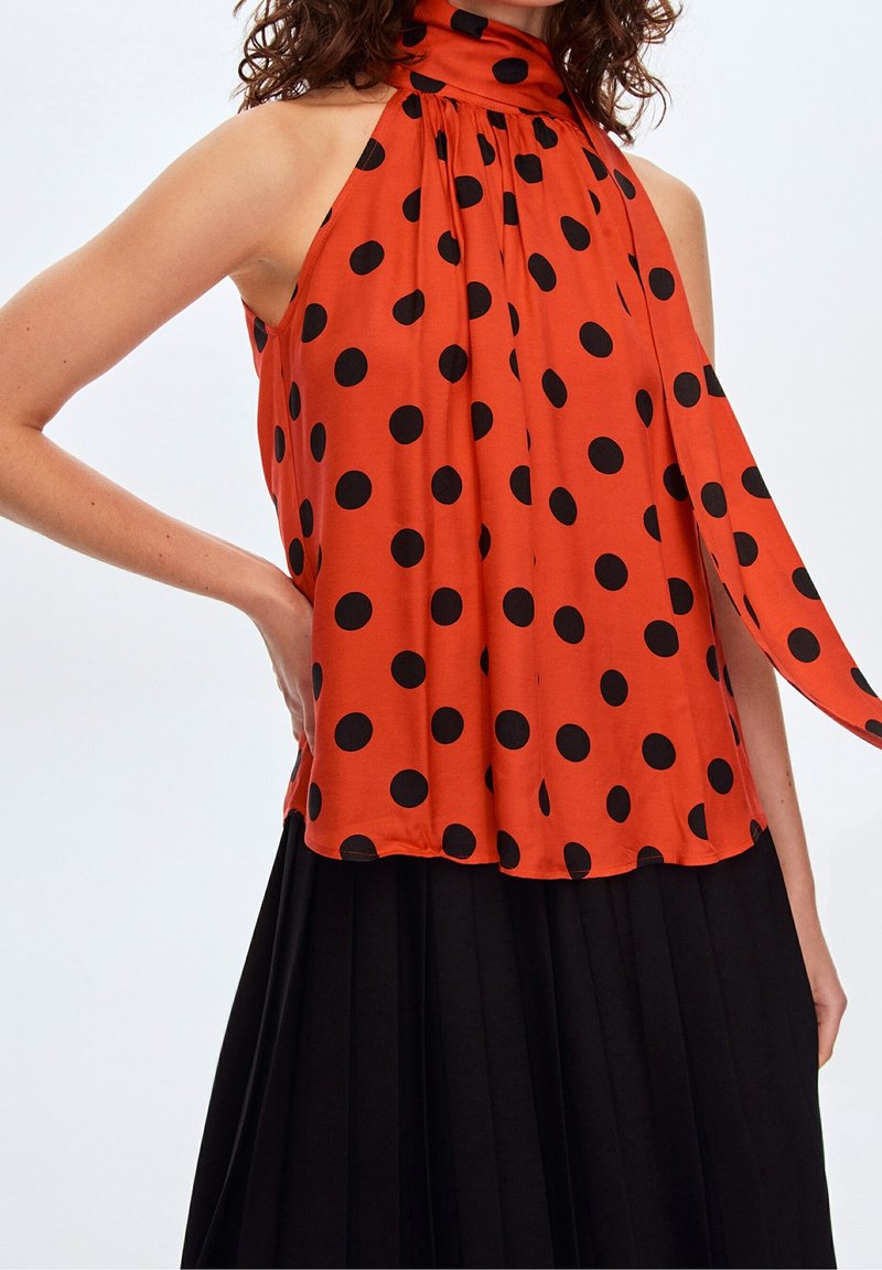 Femme portant un haut dos nu orange à pois noirs et une jupe noire plissée, main sur la hanche.